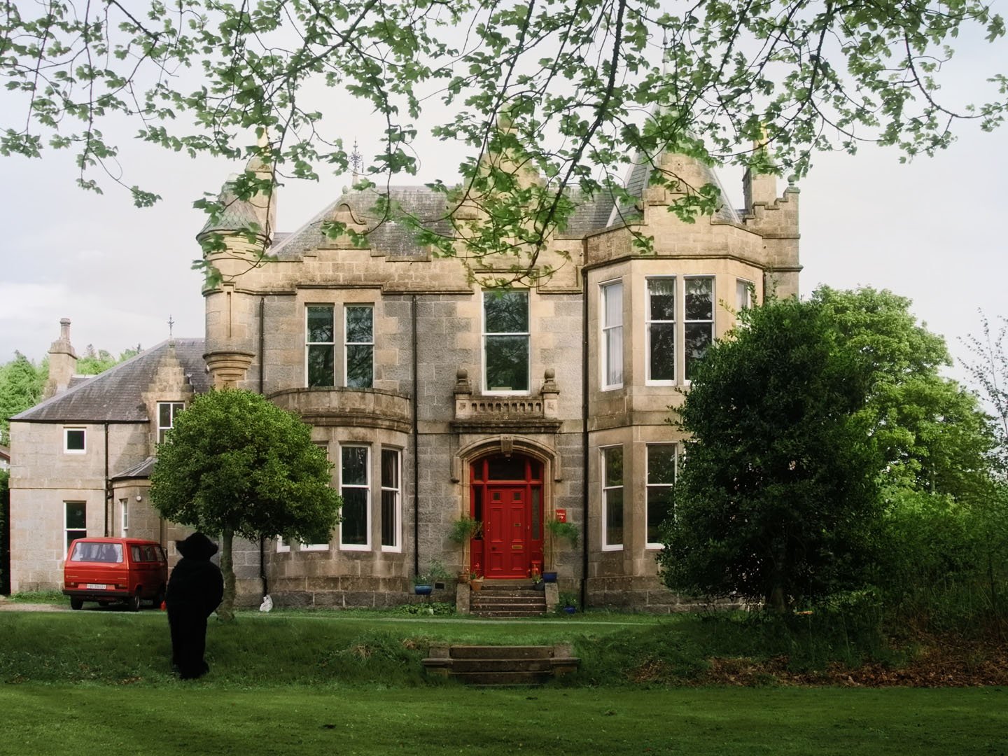 Film still: the Monster stands on a lawn facing a grand stone house with a red front door