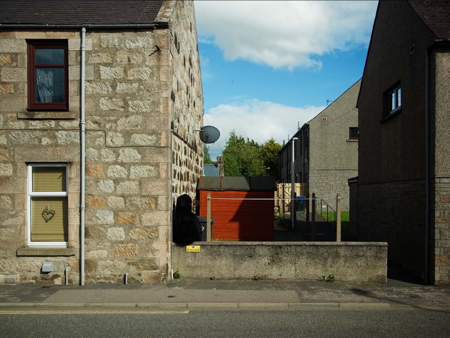 Film still: the Monster peers around the corner of a stone house near a red shed under blue sky