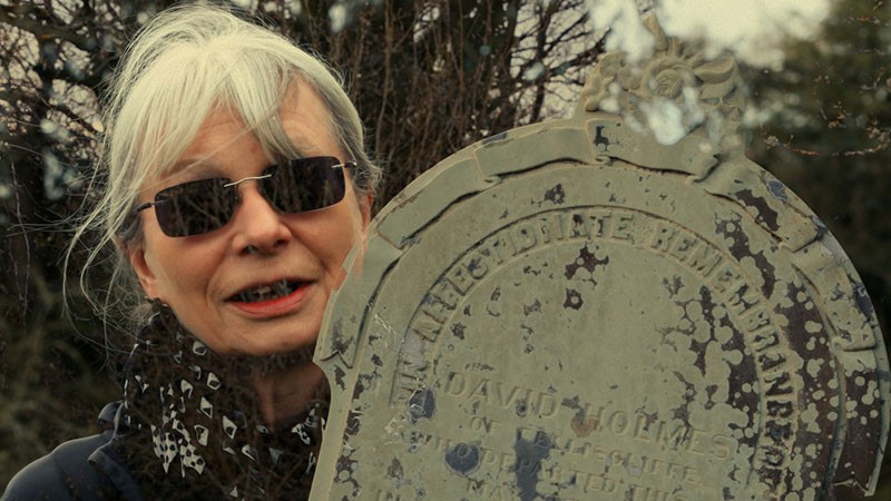 Film still: a grey-haired woman in sunglasses beside a weathered gravestone