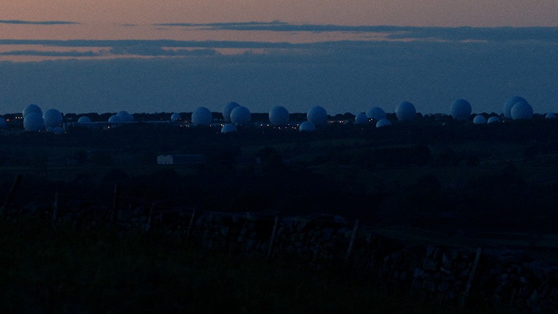 Film still: white radomes of RAF Menwith Hill on the horizon at dusk