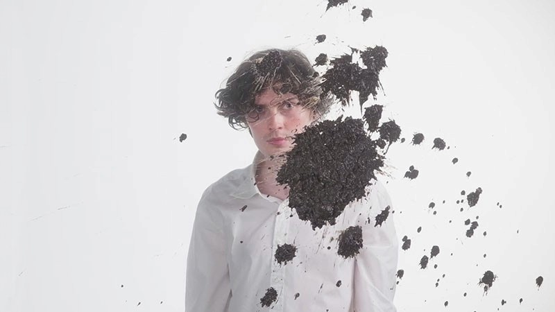 Film still: a young man in a white shirt stands against white as dark soil bursts across his chest