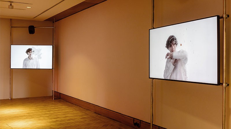 Installation view: two screens in a warm-lit gallery showing a figure in white behind spattered glass