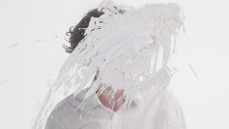 Film still: a figure's head and shoulders covered in dripping white cream against a white backdrop