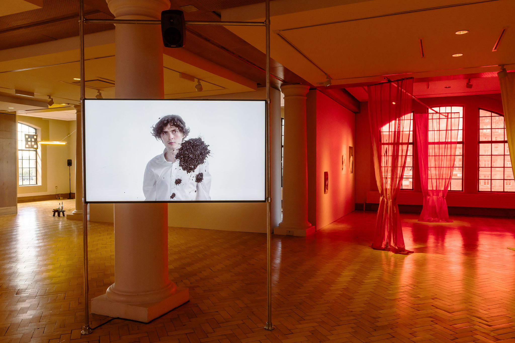 Installation view at City Art Centre, Edinburgh: screen showing a man in white as soil flies, red-lit gallery beyond