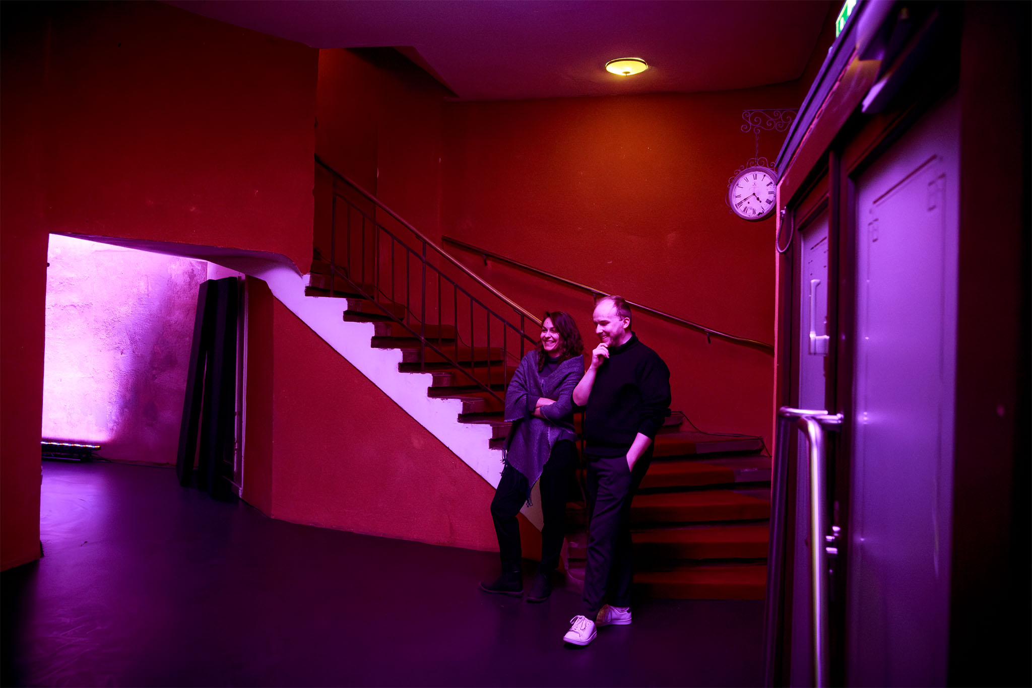 Designer Mihaela Dobreva and Daniel Hughes chat beside a staircase in the pink-lit theatre foyer