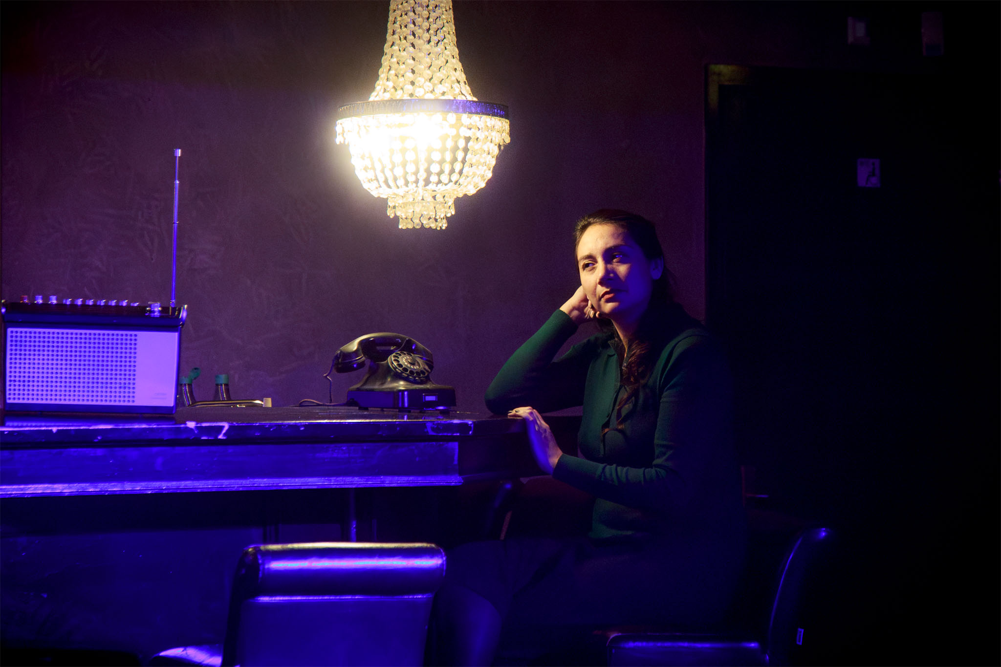 Leicy Valenzuela performs seated at the main hall bar beneath a chandelier, beside a vintage telephone and radio