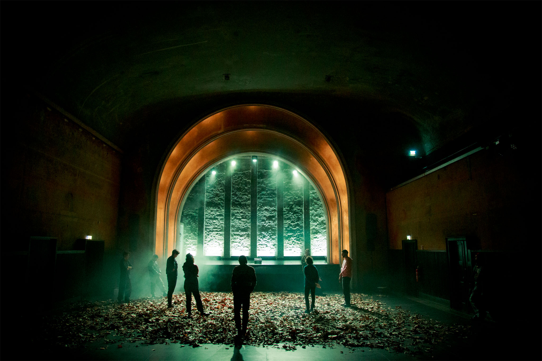 Dress rehearsal still: figures stand among Mihaela Dobreva's gathered leaves before a glowing arched window