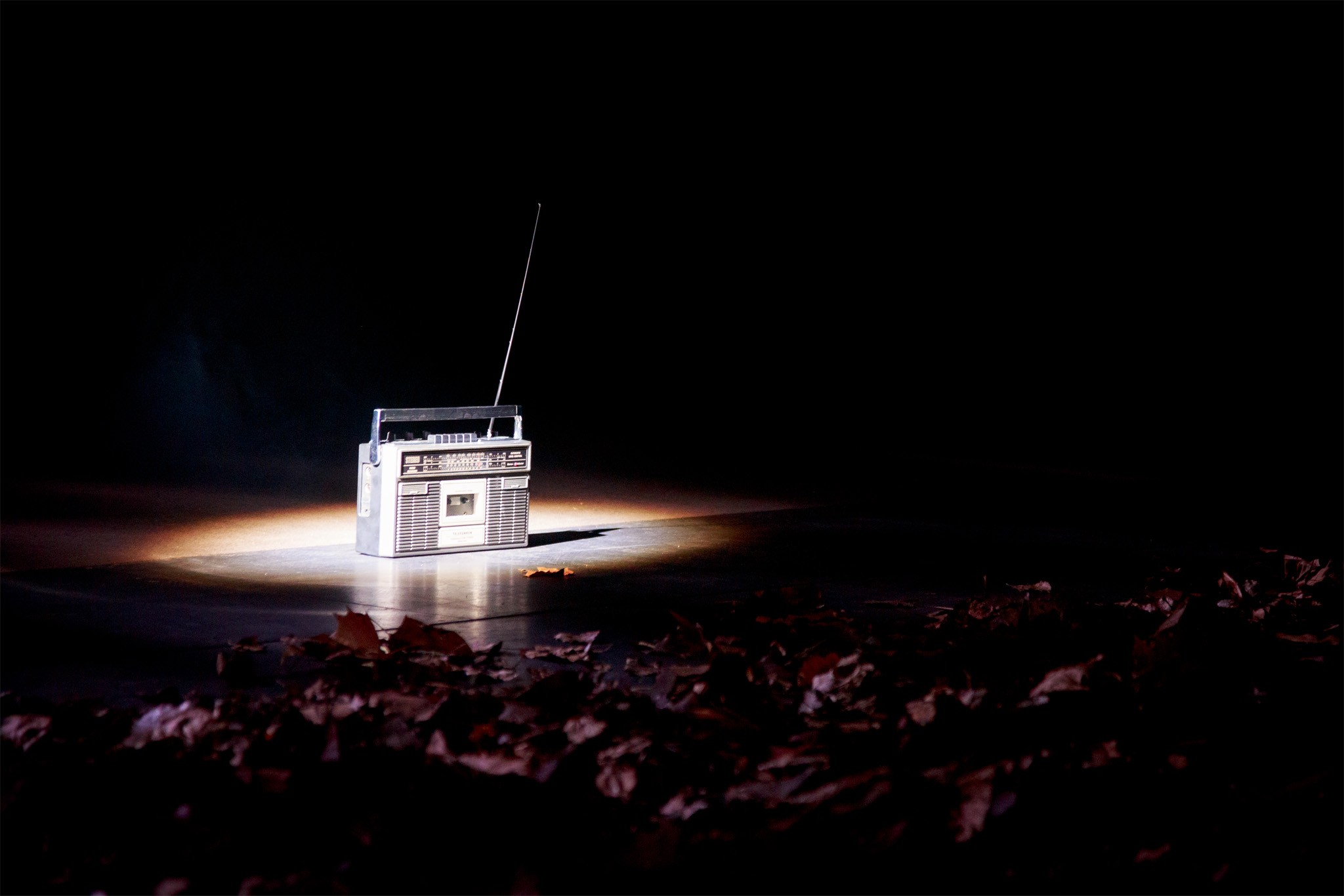 A vintage radio cassette player spotlit on the dark stage floor, autumn leaves in the foreground