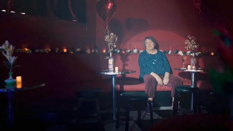 Film still: Jo Clifford sits alone among candlelit cabaret tables dotted with flowers and heart balloons