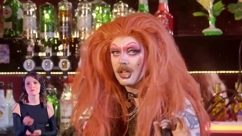 Film still: host Pink Pound, in a big red wig, tends the bar while BSL interpreter Amy Cheskin signs nearby
