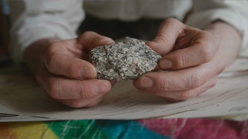 Al Baker's hands holding a speckled granite rock over papers, film still from Rocabarraigh