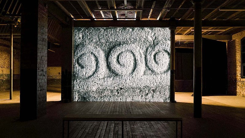 Installation view of Rocabarraigh at Market Gallery: projected spiral stone carvings facing a bench in a dark gallery