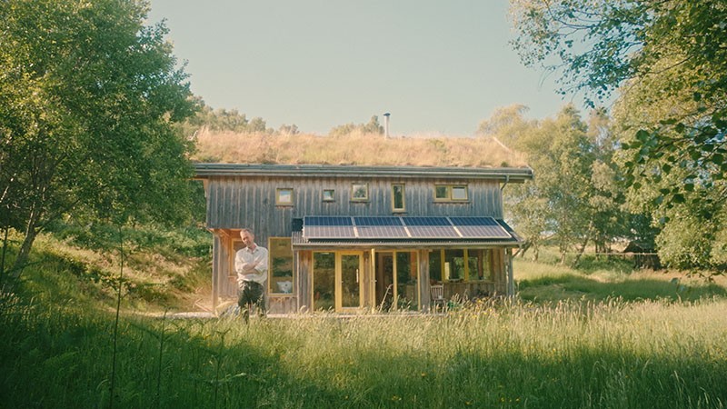 Al Baker standing outside his turf-roofed timber house with solar panels, film still from Rocabarraigh