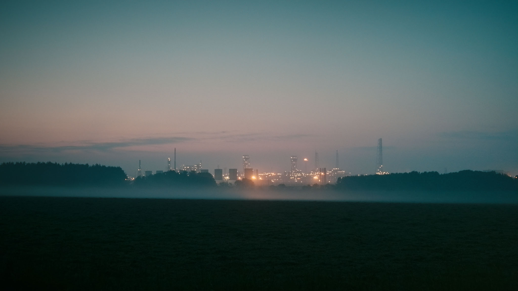 Film still of St Fergus Gas Terminal glowing at dusk beyond mist-covered fields