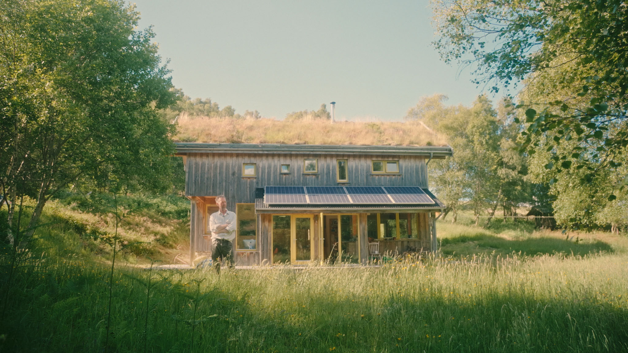Film still of Al Baker outside his turf-roofed timber house with solar panels, framed by trees and tall grass