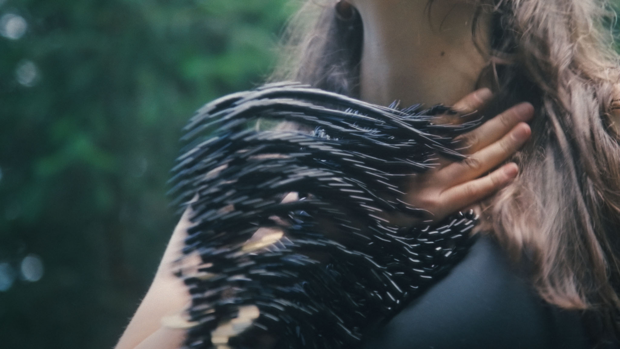 Film still close-up of Anya Sirina touching a black fringed garment at her neck, greenery blurred behind