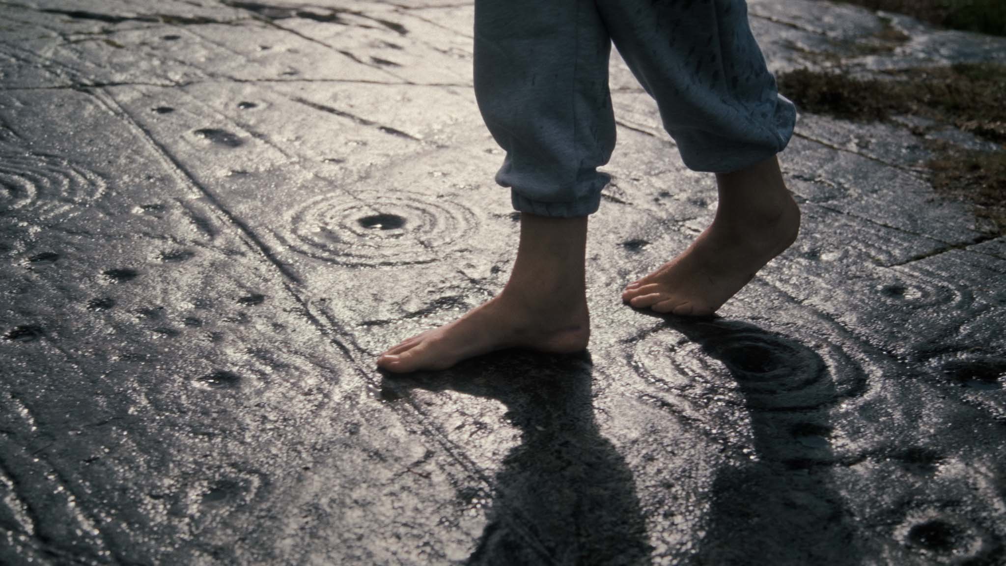 Film still of Anya Sirina's bare feet walking across wet rock carved with cup-and-ring markings