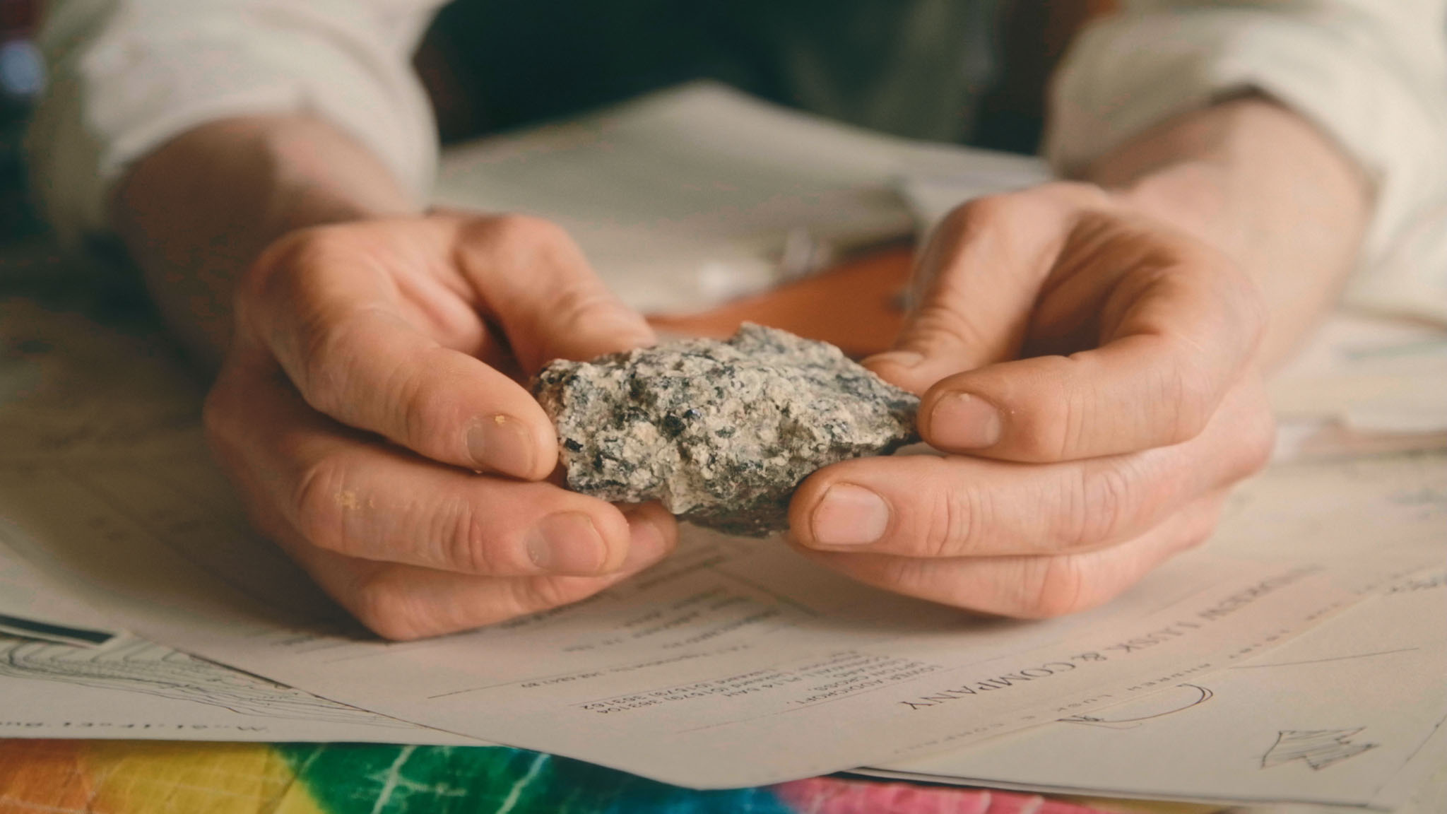 Film still close-up of Al Baker's hands holding a speckled granite rock over printed papers