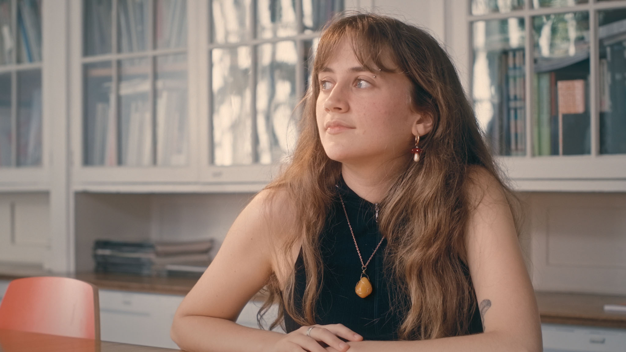 Film still of Lauren MacDonald seated before glass-fronted cabinets, wearing an amber pendant necklace