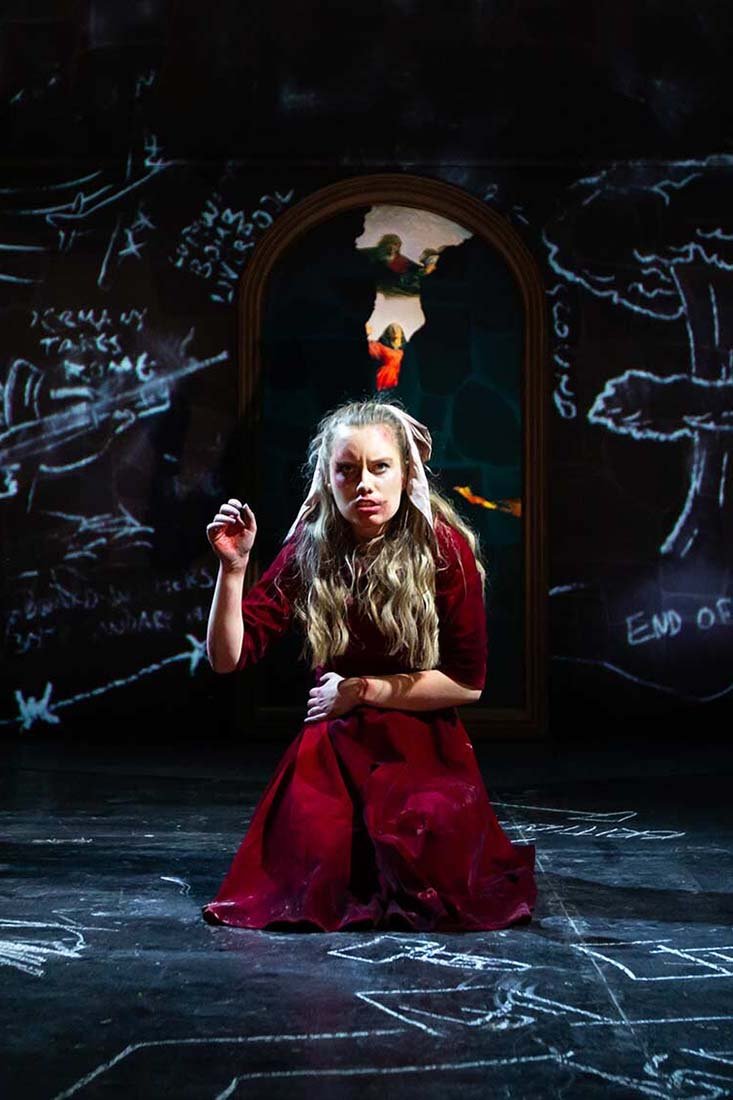 Claire Wetherall as Maria kneels in a red dress before an arched painting and chalk-covered cellar walls.