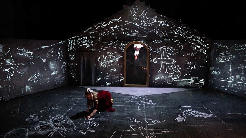 Claire Wetherall as Maria kneels chalk-drawing on the stage floor of a cellar covered in projected chalk sketches.
