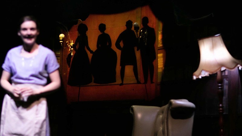 Ashley Smith as Enid in an apron before a projected puppet animation of silhouetted figures in the library.