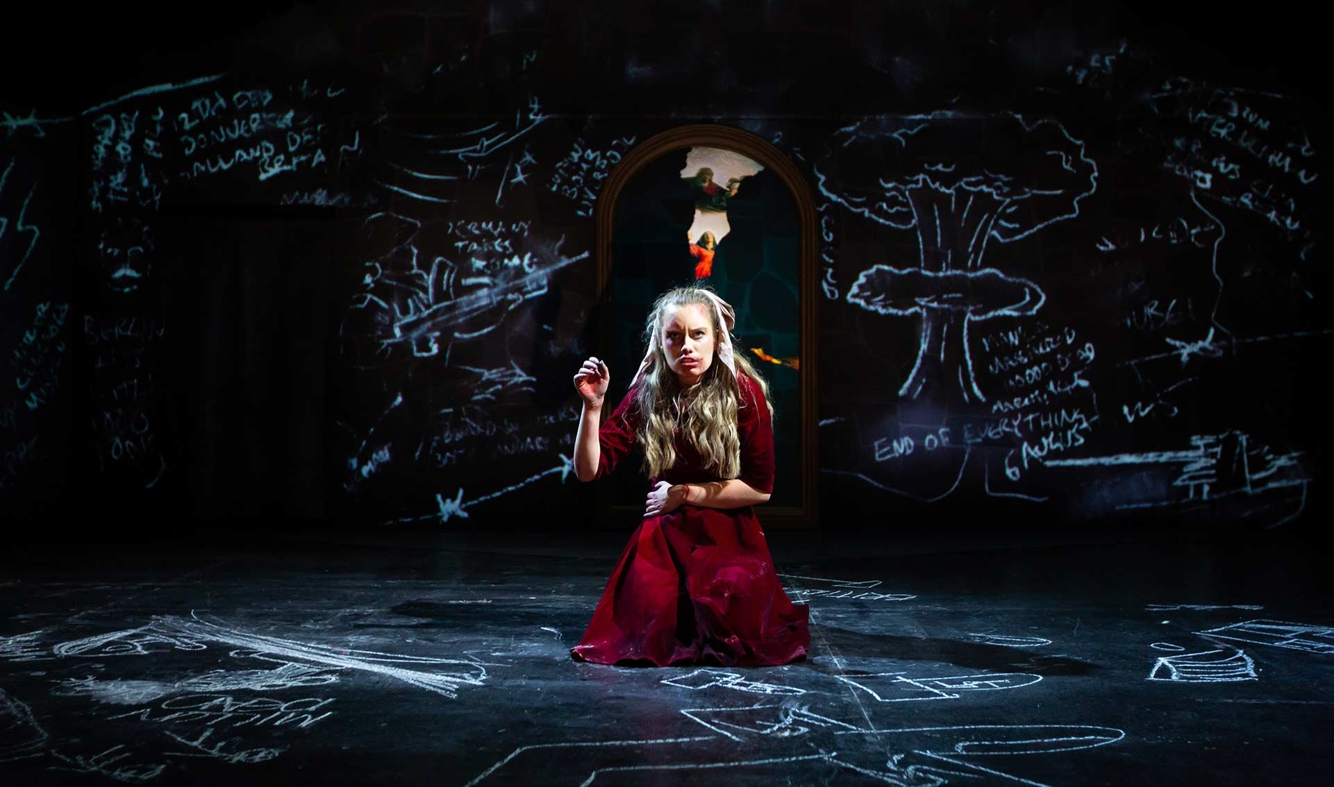 Claire Wetherall as Maria kneels amid projected chalk drawings on the cellar walls, the painting shard glowing behind her.