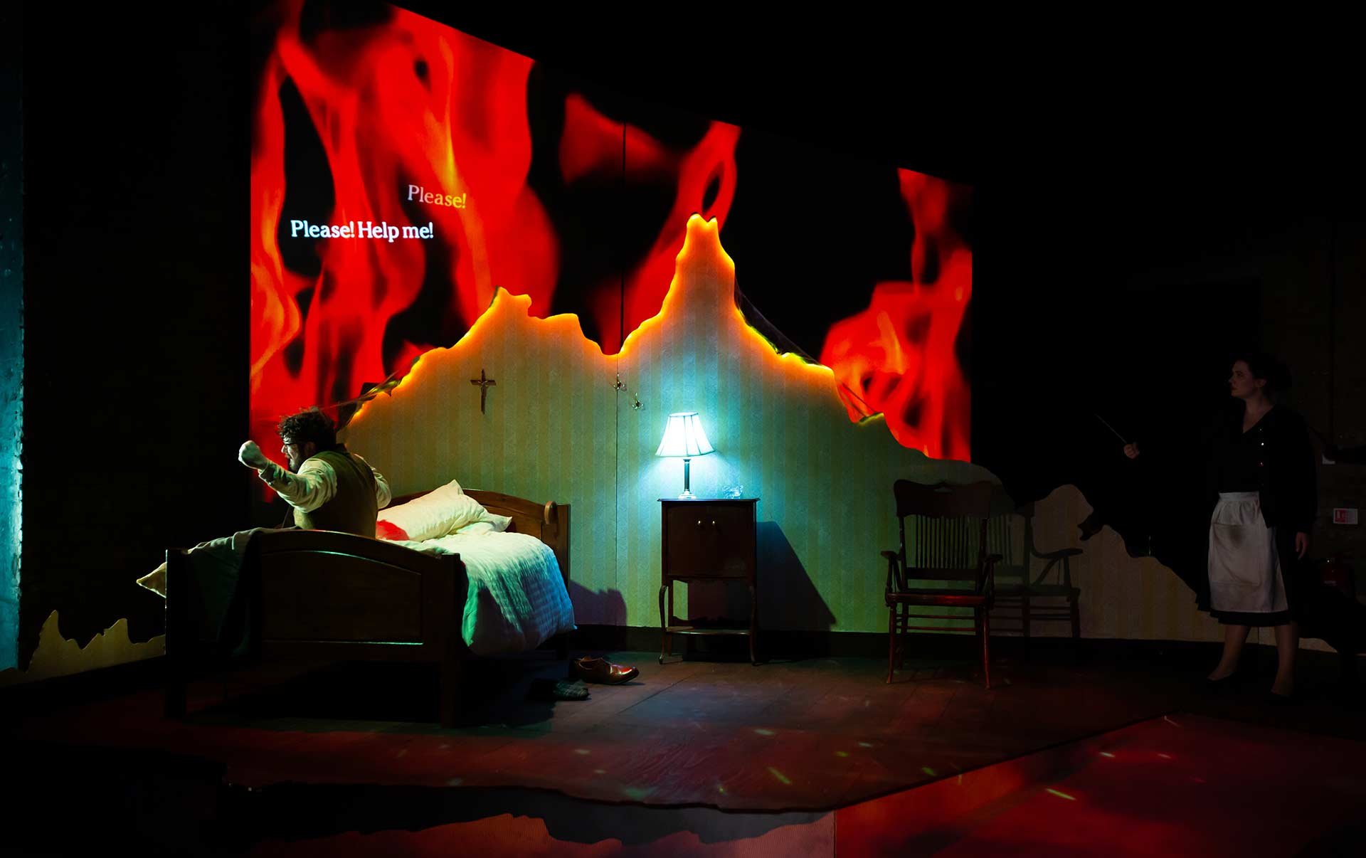 Neil Shand as Provan sits up in bed as projected flames engulf the bedroom walls; Irina Vartopeanu as Theresa looks on.