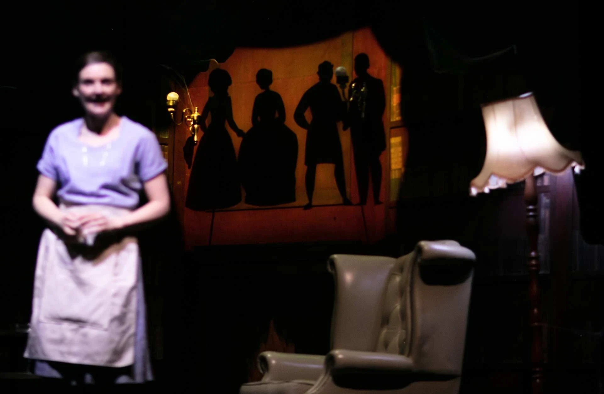Ashley Smith as Enid smiles in an apron while silhouetted puppet figures are projected in the library behind her.