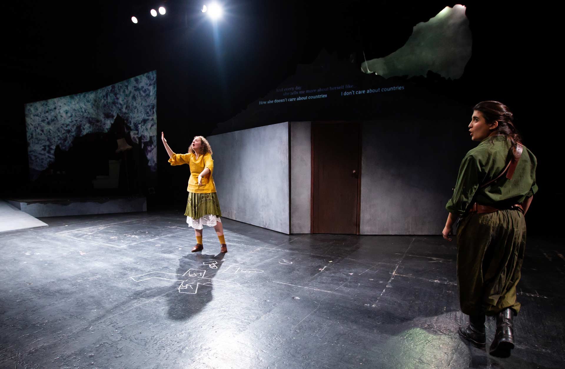 Amy Helena as Queenie gestures to Benedetta Zanetti as Mama K, with a sea-cliff projection and captions above the set.