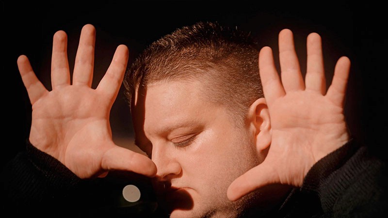 Jamie Black, eyes closed, raising open palms beside his face in warm light, Tunnel Blue music video still